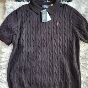 Brun kabelstickad pikétröja från Polo Ralph Lauren - Säljer en brun kabelstickad pikétröja från Polo Ralph Lauren med klassisk krage och korta ärmar. Tröjan har ett broderat rött logomärke på bröstet och är tillverkad i mjukt material. Perfekt för en stilren look.