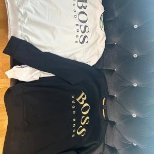 Svart och vit sweatshirt från Hugo Boss - Två stilrena sweatshirts från Hugo Boss, en svart med guldig logga och en vit med svart logga. Båda har rund halsringning och lång ärm. Perfekta för en snygg och enkel look.
