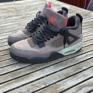 Nike Air Jordans 4 Taupe Haze - Säljer ett par Nike Air Jordans 4 i färgen Taupe Haze med svarta och röda detaljer. Skorna har svart snörning, meshpaneler och en synlig Air-sula. Insidan är fodrad i rött och de har en klassisk Jumpman-logga på plösen och hälen.