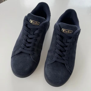 Svarta sneakers från Polo Ralph Lauren - Snygga svarta sneakers från Polo Ralph Lauren med ovandel i mocka och klassisk snörning. Skorna har rund tå och platt sula, samt diskret Polo-logga på plösen och sidan. Perfekta för en stilren vardagslook. Den är använda ett få antal gånger. Hör av er vid intresse