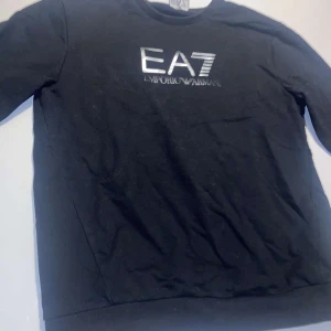 Svart hoodie från Emporio Armani EA7 - Säljer en svart hoodie från Emporio Armani EA7 med stor logga i silver på bröstet. Tröjan har långa ärmar, huva och ribbade muddar. Perfekt för en stilren och sportig look. Använd 1-5 gånger. 