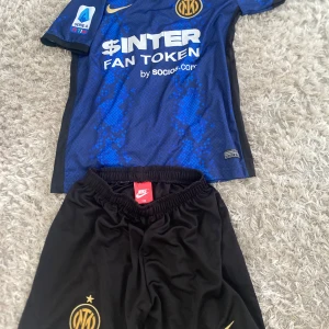 Inter Milan matchtröja och shorts Nike - Säljer en blå och svart Inter Milan fotbollströja från Nike med guldiga detaljer och klubbmärke, samt tillhörande svarta shorts med guldiga loggor. Tröjan har korta ärmar och ett mönster i blått och svart. Perfekt för fotbollsträning eller match.