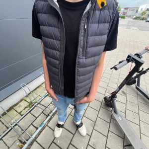 Svart dunväst från parajumper - Säljer en svart dunväst från para jumper  med dragkedja framtill och ståkrage. Västen har en quiltad design och en liten orange detalj vid axeln. Perfekt att slänga över en hoodie eller t-shirt för en cool look. Storlek xs men passar s