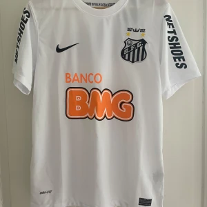 Santos FC fotbollströja från 12-13 - Säljer en vit Santos FC matchtröja från Nike med svarta detaljer och tryck. Tröjan har klubbmärke på bröstet, sponsorloggor (BMG, NETSHOES) och Dri-Fit-material för bra ventilation. Perfekt till sommaren