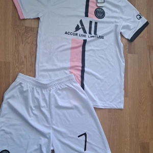 PSG Mbappé 7 fotbollsdress - Vit PSG fotbollsdress med rosa och svart vertikal rand, Mbappé och nummer 7 på ryggen. Kortärmad modell med rosa detaljer på ärmsluten och klubbmärke på bröstet. Helt ny me etikettet på och påsen