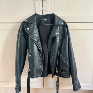 Svart skinnjacka från Missguided - Säljer en svart skinnjacka från Missguided med klassisk bikerstil. Jackan har silvriga dragkedjor, snedställda fickor, bälte i midjan och långa ärmar med dragkedjor vid ärmsluten. Passformen är mer åt oversize boxig. Storlek 32,4, men jag skulle säga den är stor i storleken, och därför har varit oversize för mig som är 160cm och bär s/m i kläder