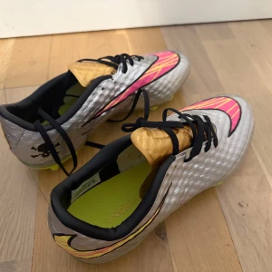Nike Hypervenom fotbollsskor silver/gul - Säljer ett par Nike Hypervenom fotbollsskor i silver med gula och rosa detaljer. Skorna har svart snörning, rund tå och platt sula med dobbar för gräsplan. Swoosh-logga på sidan och dödskalletryck på hälen. Perfekta för dig som vill sticka ut på planen.