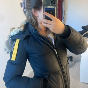 Svart vinterjacka från Parajumpers - Säljer en svart lång vinterjacka från Parajumpers i storlek XSS. Jackan har dragkedja, knappar och två stora fickor framtill samt en gul detalj på ärmen. Perfekt för kalla vinterdagar. Den är bara använd en vinter. Den är i bra skick!