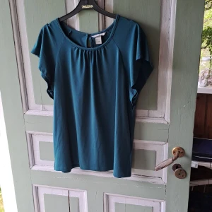 Petrolblå blus med kort ärm - Säljer en enkel och stilren petrolblå blus med rund halsringning och korta, lösa ärmar. Blusen har ett litet sprund med knapp i nacken och är tillverkad i ett mjukt, slätt material. Perfekt för en avslappnad men snygg look.