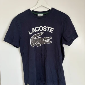 Mörkblå t-shirt från Lacoste - Säljer en mörkblå t-shirt från Lacoste med stor vit logga och krokodiltryck på bröstet. T-shirten har rund halsringning och normal passform. Perfekt för en avslappnad och sportig stil.