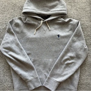 Grå hoodie från Polo Ralph Lauren - Klassisk grå hoodie från Polo Ralph Lauren med broderad blå logga på bröstet. Tröjan har huva med dragsko och en stor magficka framtill. Perfekt för en avslappnad stil.