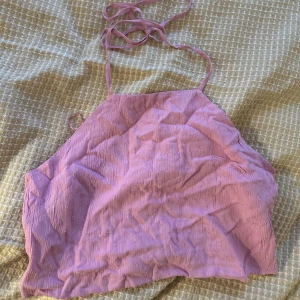 Ljusrosa croppad halterneck topp - Säljer en ljusrosa croppad halterneck-linnetopp med knytband i nacken. Toppen har en lätt strukturerad yta och är perfekt för varma dagar. Enkel och stilren design som passar till mycket. Använd endast en gång