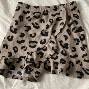 Beige omlottshorts med leopardmönster från 157 - Säljer ett par beige kjol med svart leopardmönster från 157. Kjolen har volangdetalj längs kanten och är gjorda i ett lätt, strukturerat tyg. Perfekta för en trendig och avslappnad look.