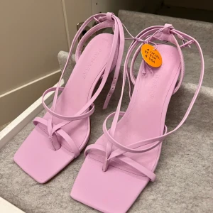 Ljuslila/rosa sandaler med snörning - Säljer ett par ljuslila sandaler med fyrkantig tå och tunna remmar som knyts runt vristen. Skorna har en stilren design och passar perfekt till vår och sommar. Storlek 37. 