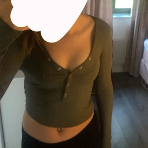 Grön croppad ribbad topp med knappar - Snygg grön croppad topp med lång ärm och djup v-ringning. Toppen har ribbad struktur och knappar framtill, perfekt för en trendig vardagslook.