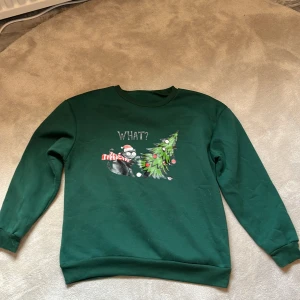 Grön jultröja med tryck - Mörkgrön sweatshirt med lång ärm och rund hals. Framsidan har ett tryck med en katt i tomteluva, en vält julgran och texten 'WHAT?'. Perfekt för julens alla tillfällen och för dig som gillar roliga motiv. Vet inte vart den kommer ifrån! Hör av er vid frågor eller intresse❤️