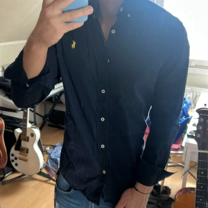 Mörkblå skjorta från Ralph Lauren - Säljer en klassisk mörkblå skjorta från Ralph Lauren med gul broderad logga på bröstet. Skjortan har vita knappar och lång ärm, perfekt för en stilren look. Passformen är normal och materialet känns som bomull.