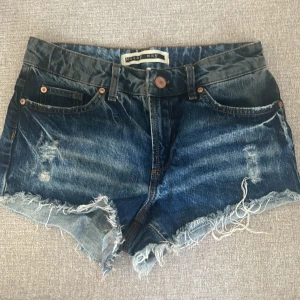 Blå jeansshorts från Noisy May - Säljer ett par blå jeansshorts från Noisy May med slitna detaljer och fransig kant. Klassisk femficksmodell med knapp och dragkedja framtill. Perfekta för varma dagar och en avslappnad stil.