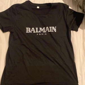 Svart t-shirt från Balmain Paris - Snygg svart t-shirt från Balmain Paris med vit logga tryckt på bröstet. Klassisk rund halsringning och korta ärmar. Perfekt för dig som gillar stilrena och trendiga plagg.