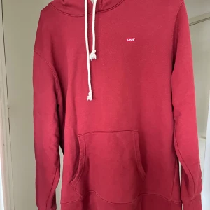 Röd hoodie från Levi's - Säljer en röd hoodie från Levi's i storlek M. Tröjan har en klassisk känguruficka framtill, vit dragsko i huvan och liten Levi's-logga på bröstet. Perfekt för en avslappnad stil.
