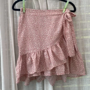 Rosa omlottkjol med volang - Supersöt rosa omlottkjol med vitt leopardmönster och volangkant. Kjolen har knyt i sidan och är i en luftig, lätt kvalitet som passar perfekt till varma dagar. 48 cm lång, perfekt för sommaren. Resor i midjan.