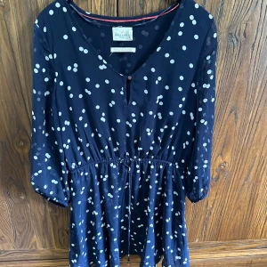 Mörkblå prickig klänning/bluse  - Supersöt mörkblå klänning med vita prickar. Klänningen har v-ringning, knappar framtill, trekvartsärmar och dragsko i midjan för en snygg passform. Perfekt för dig som gillar lekfulla mönster och bekväma plagg.