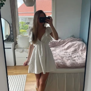 Vit kortklänning med volangärmar och v-ringning - Superfin vit klänning med djup v-ringning och volangdetaljer på axlarna. Klänningen är kort och har en luftig passform, perfekt för varma dagar. Snygg och enkel design som känns både trendig och söt.