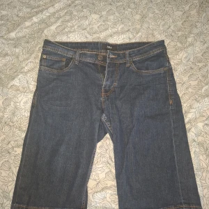Mörkblå jeansbyxor från Smog - Säljer ett par klassiska mörkblå jeans från Smog i storlek M. Byxorna har fem fickor, orangea sömmar och normal passform. Perfekta för dig som gillar en enkel och tidlös stil.
