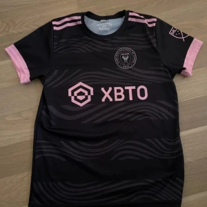 Inter Miami CF Messi 10 fotbollströja - Säljer en svart och rosa Inter Miami CF fotbollströja med Messi och nummer 10 på ryggen. Tröjan har korta ärmar, rosa detaljer och klubbmärke samt sponsorlogga på bröstet. Perfekt för fans av Messi och MLS. Passar ungefär 158-174
