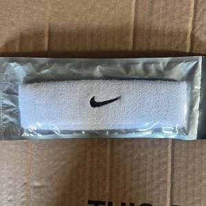 Vit headband från Nike - Säljer ett vitt headband från Nike med klassisk svart logga broderad framtill. Perfekt för att hålla håret på plats under träning eller sport. Materialet är mjukt och absorberande. Aldrig andvänt den! Den är fortfarande i förpackningen 