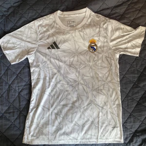 Vit Real Madrid fotbollströja från Adidas - Säljer en vit Real Madrid fotbollströja från Adidas med grått grafiskt mönster och klubbmärke på bröstet. Tröjan är kortärmad och har en modern, sportig design. Perfekt för fans eller träning.