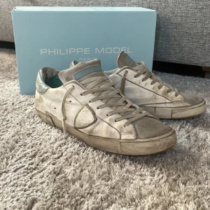 Vita sneakers från Philippe Model - Snygga vita sneakers från Philippe Model med grå detaljer och ljusblå inslag på plösen och hälen. Skorna har snörning, rund tå och platt sula. Perfekta för en stilren och avslappnad look.