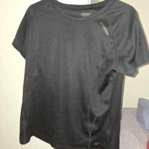 Svart tränings-t-shirt från Hummel - Svart sportig t-shirt från Hummel med diskret logga på bröstet och klassiska pilar på ärmen. Tillverkad i ett lätt och ventilerande syntetmaterial, perfekt för träning eller fotboll. Normal passform och korta ärmar.