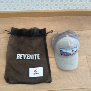 Grå limited edition mocka keps från revenite - Snygg grå keps från Revenite med ett färgstarkt tryck framtill som föreställer en kvinna vid en röd sportbil och en yacht i bakgrunden. Kepsen har mesh baktill för extra ventilation och justerbar passform. Limited edition och går ej att köpa igen. Finns en liten fläck i kepsen(se sista bild) pris går självklart att diskutera. Mvh Levi