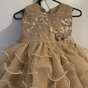 Champagnefärgad festklänning med volanger - Säljer en superfin champagnefärgad klänning med broderade blommor upptill och volangkjol i flera lager. Klänningen har ärmlös design och en söt rosettdetalj i midjan. Perfekt för speciella tillfällen!