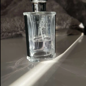 Prada L'Homme parfym - Elegant och stilren parfymflaska från Prada med en transparent design och svarta detaljer på sidorna. Flaskan har en metallisk kork och rymmer en doft som är både modern och tidlös. Perfekt för den som vill ha en sofistikerad och fräsch doft.