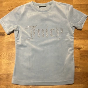 Ljusblå velour t-shirt från Juicy Couture - Säljer en ljusblå t-shirt i mjuk velour från Juicy Couture med glittrande stenar som bildar texten 'Juicy' på bröstet. Nästan aldrig använt! T-shirten har rund hals och korta ärmar, perfekt för en trendig och avslappnad look.