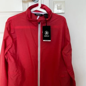 Röd vindjacka från Abacus - Säljer en snygg röd vindjacka från Abacus med dragkedja framtill. Jackan har en hög krage och är perfekt för blåsiga dagar. Den är lätt och smidig att bära med sig. Passar bra för utomhusaktiviteter och håller dig skyddad mot vinden. Helt ny, prislapp kvar. Nypris: 900 kr