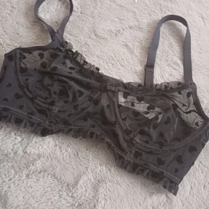 Svart hjärtmönstrad bh - Säljer en svart korsett med hjärtmönster och justerbara axelband. Den har en söt volangdetalj längs kanterna och stängs med hyskor i ryggen. Perfekt för en romantisk look.