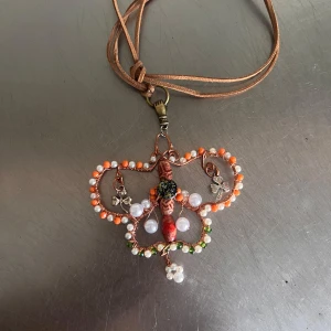 Handgjort halsband med fjärilshänge och pärlor - Unikt halsband med ett stort fjärilshänge dekorerat med orange, vita och gröna pärlor samt små metallfjärilar. Hänget har inslag av koppartråd och hänger i ett brunt läderband. Perfekt för dig som gillar bohemisk stil.