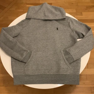 Grå hoodie från Polo Ralph Lauren - Säljer en stilren grå hoodie från Polo Ralph Lauren med klassisk svart logga broderad på bröstet. Tröjan har huva, känguruficka och ribbade muddar. Perfekt för en avslappnad look.