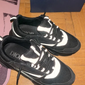 Svarta och vita sneakers från Dior - Säljer ett par snygga svarta och vita sneakers från Dior med coola detaljer och tjock sula. Skorna har snörning och en sportig design med mesh och syntetmaterial. Perfekta för dig som vill sticka ut med stilrena och lyxiga sneakers.