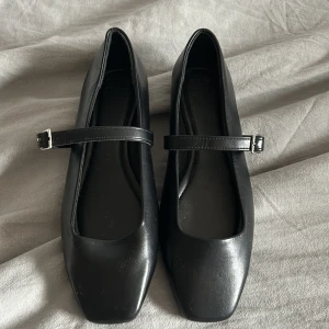 Svarta  ballerinaskor med spänne - Säljer ett par svarta Mary Jane ballerinaskor från ASOS med fyrkantig tå och justerbart spänne över foten. Skorna har platt sula och är tillverkade i syntetmaterial. Perfekta för en stilren och klassisk look.