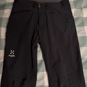 Haglöfs byxor - Svarta täckbyxor från Haglöfs med dragkedjeförsedda fickor och logga på vänster ben. Byxorna har normal passform och är perfekta för kalla dagar utomhus. Stängs med knappar och dragkedja framtill. Fraktas endast!