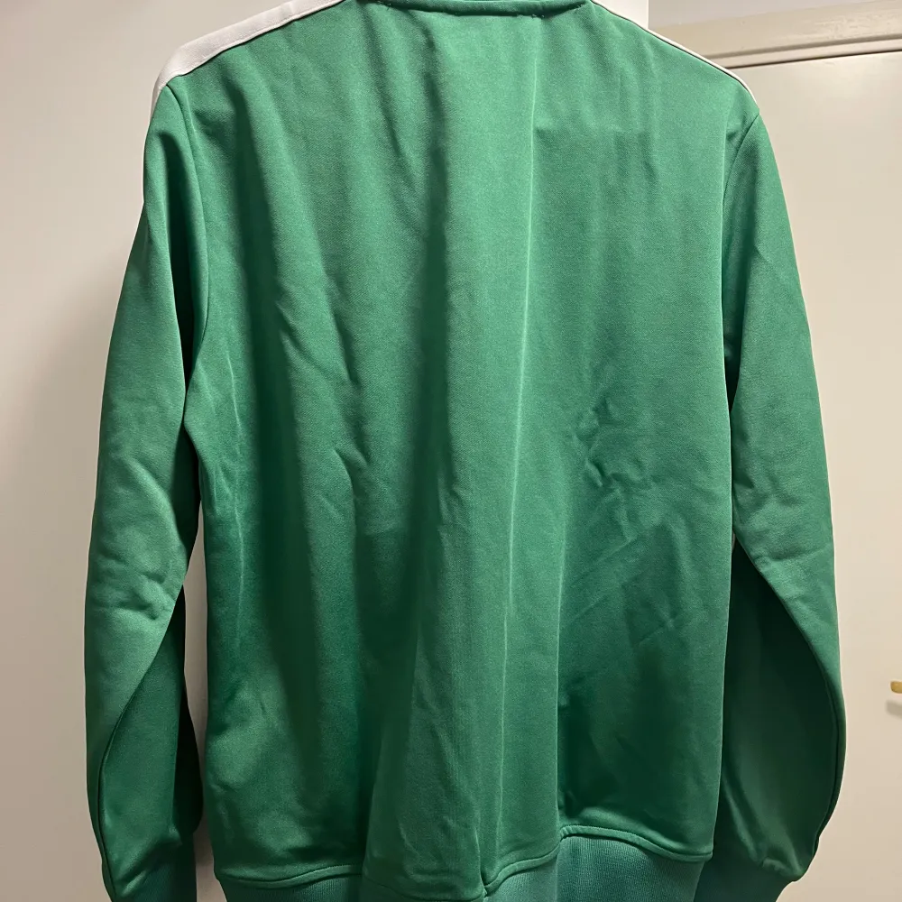 Säljer en grön track jacket från Palm Angels med vit dragkedja och vita ränder längs ärmarna. Jackan har hög krage, två fickor med dragkedja och logga tryckt på bröstet. Perfekt för en sportig och trendig look. Helt oanvänd!. Takit.