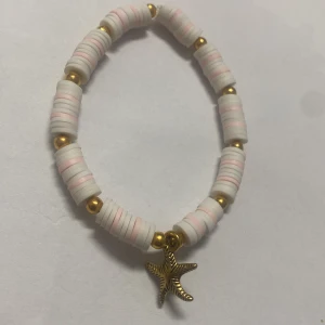 Armband med vita och rosa pärlor - Charmigt armband med vita och rosa platta pärlor samt guldfärgade kulor. Armbandet har olika hängen, inklusive en sjöstjärna, en skylt med texten 'Buena Suerte' och ett roder. Perfekt för att ge en personlig touch till din stil.