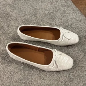 Vita ballerinaskor med rosett från NA-KD - Säljer ett par vita ballerinaskor från NA-KD med flätad struktur och söt rosett framtill. Skorna har fyrkantig tå och platt sula, storlek 40