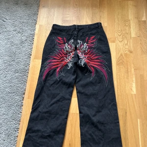 Svarta jeans med röd broderad design - Unika svarta jeans med bred passform och stor röd och vit broderad design på baksidan. Jeansen har klassisk femficksmodell och raka ben. Perfekta för dig som vill sticka ut med en cool detalj på byxorna.