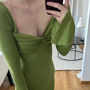 Grön långärmad kortklänning med twistad detalj - Säljer en snygg grön kortklänning med långärmade trumpetärmar och twistad detalj vid bysten. Klänningen har djup v-ringning och figurnära passform. Perfekt för dig som vill sticka ut med en trendig look!