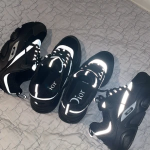 Svarta Dior sneakers med reflexdetaljer - Säljer ett par svarta Dior sneakers med tydliga vita reflexdetaljer och snörning. Skorna har en chunky sula och märkets logga syns både på sidan och på plösen. Perfekta för dig som vill sticka ut med stilrena men ändå unika sneakers.